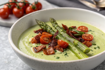 White bowl of creamy velouté d'asperges blancs garnished with fresh asparagus tips and microgreens on light gray granite countertop.